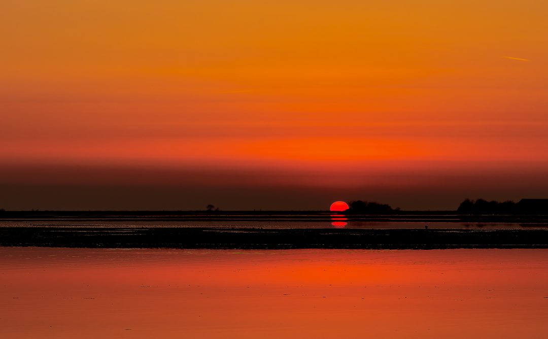 Tramonto rosso sulla laguna di Grado: il sole tocca l’orizzonte dritto, spostato verso destra, e si riflette sull’acqua.