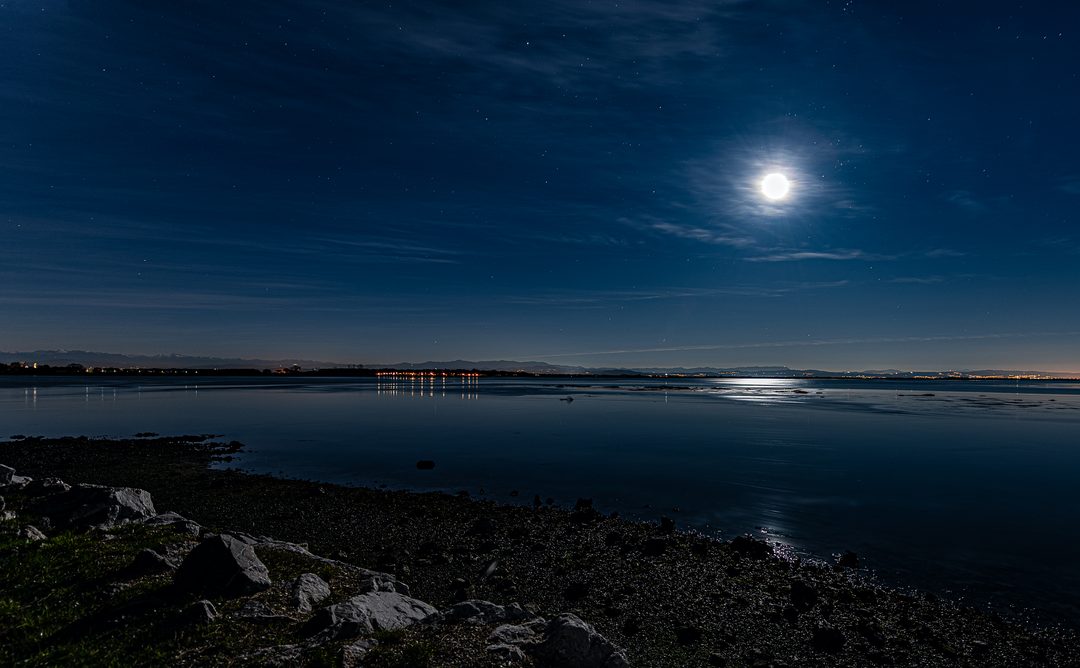 Notte sulla laguna di Grado: la luna con l’alone, le stelle, gli scogli in primo piano e le luci lontane sull’acqua ferma.