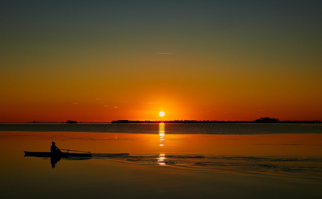 Sunset on the Grado lagoon: the sun low on the horizon and the silhouette of a person in a boat on the golden water.