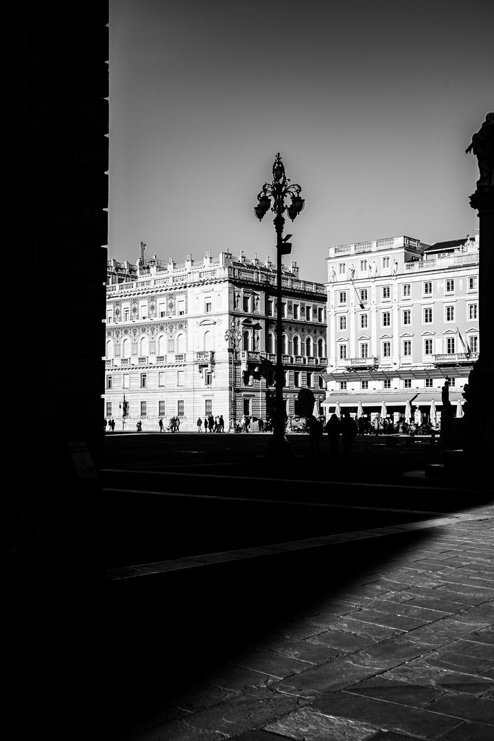 Black and white: Piazza Unità d’Italia seen from the shade, with a dark lamp post in front of the sunlit buildings and people crossing the square.